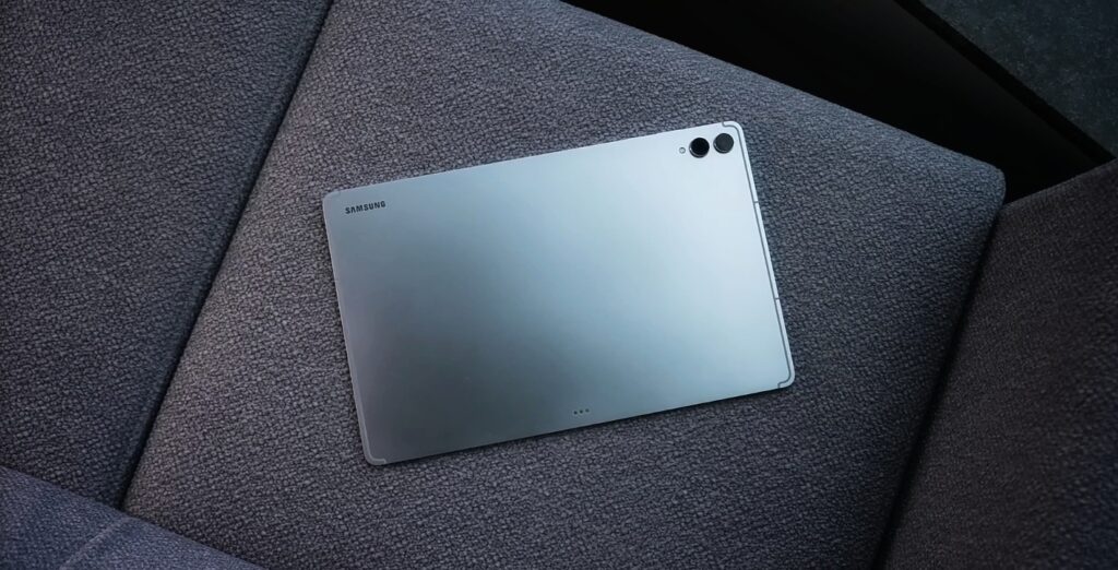 A silver Samsung Galaxy Tab S11 ultra tablet rests on a gray fabric surface, with the Samsung logo visible on it's back. The tablet features a dual-camera system and a flash in the top left corner.