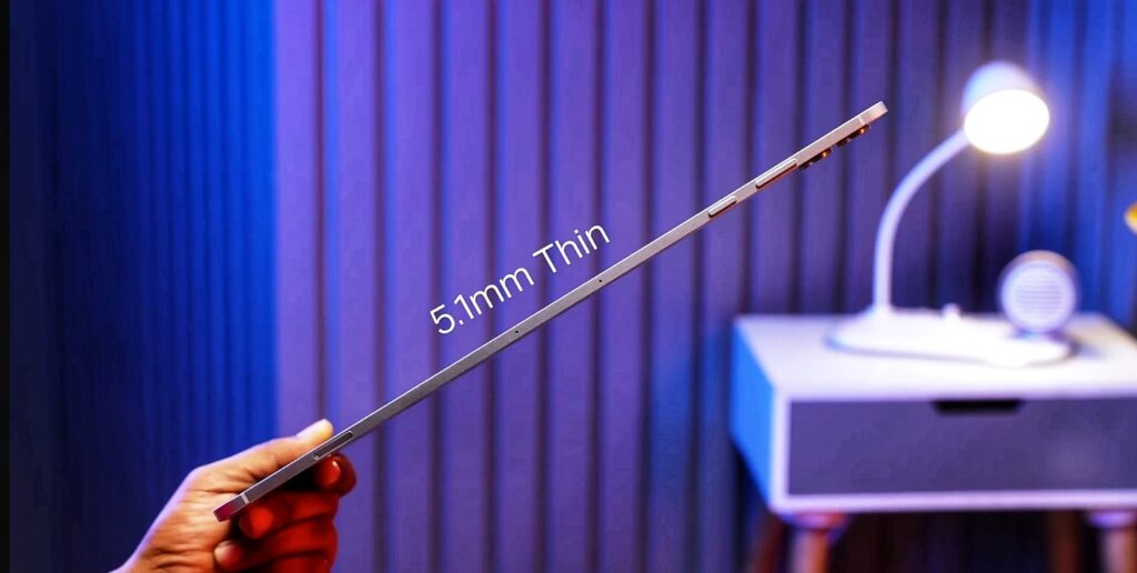 A hand holds a very thin electronic tablet or laptop, with the text “5.1mm thin" overlaid on the image to highlight it's slim profile. The device is shown from the side, with a blurred background of a room containing a desk lamp and a nightstand